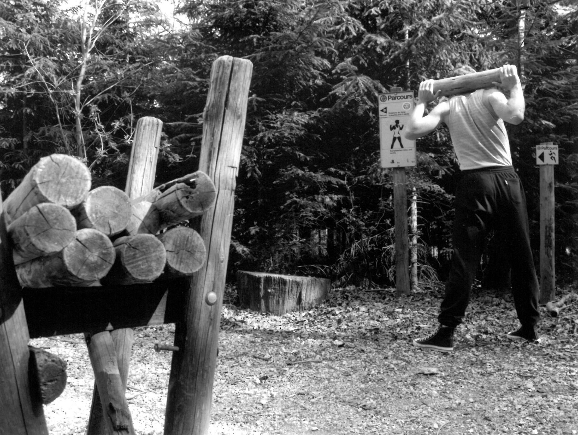 An image shows a person in sportswear carrying a heavy log on their shoulders. The person is outdoors in a wooded area, standing next to a pile of logs.