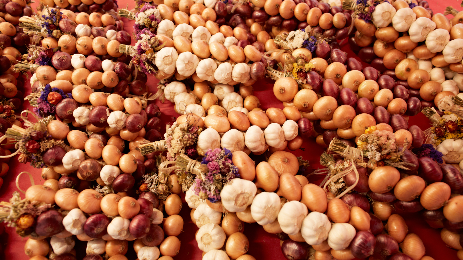 Garlands of onions and garlic in different colors, decorated with dried flowers, laid out on a red surface.