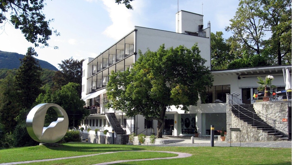 Modern multi-storey building with balconies, surrounded by trees, featuring a circular metal sculpture in the garden at the front.