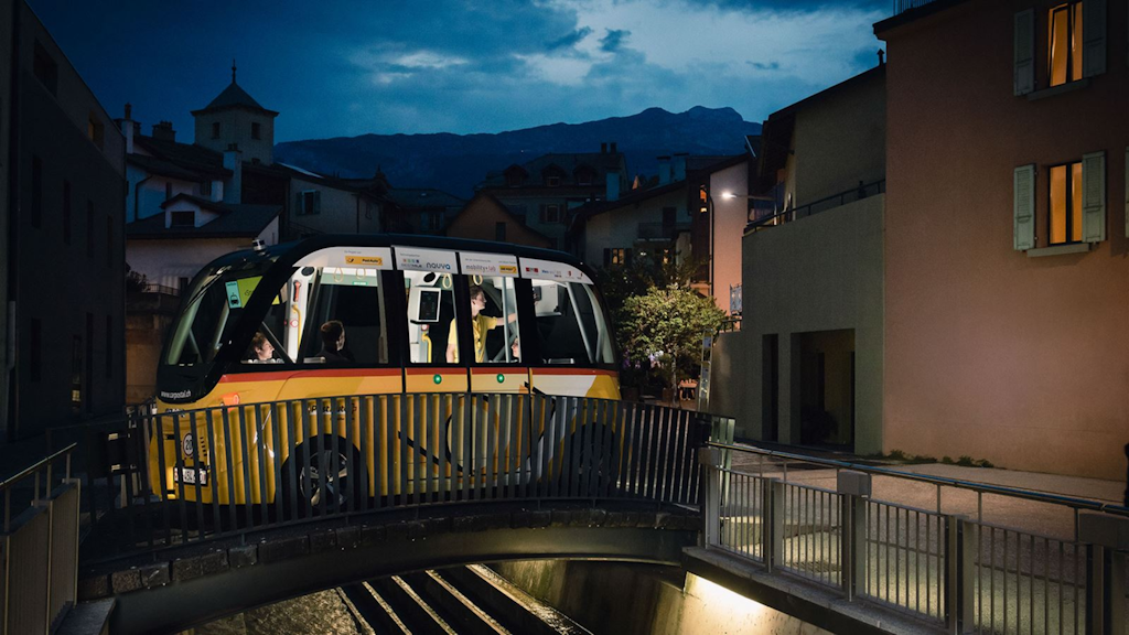 Driverless yellow PostAuto shuttle crossing a small urban bridge at night, lit interior with passengers, buildings’ façades and mountains in the background.