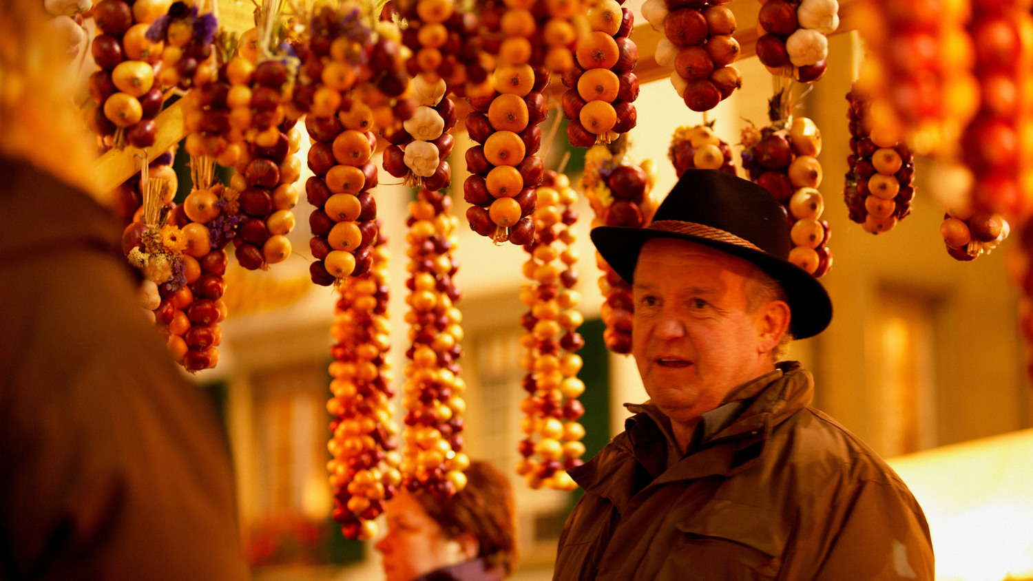 Man with a hat at a market, surrounded by hanging garlands of colorful onions and garlic, with a bright, blurred background.