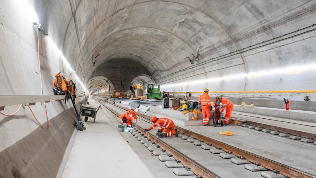 Workers in orange protective clothing installing railway tracks inside a rail tunnel under construction, with tools, machinery and materials along the sides.