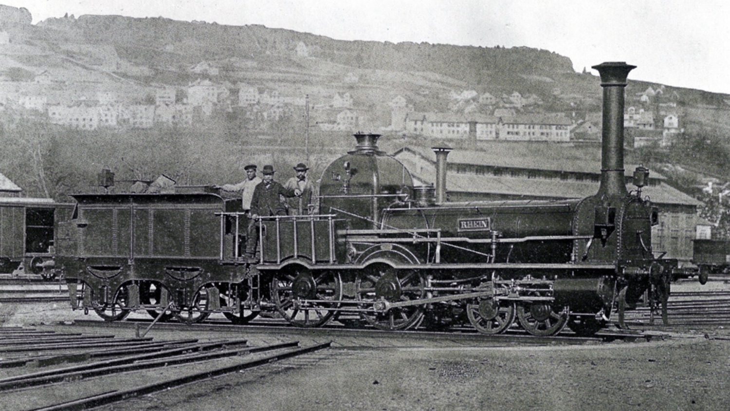 Vintage photograph of the 1867 steam locomotive ‘Rhein’ with its tender, standing on tracks before an engine shed and a hillside beyond.