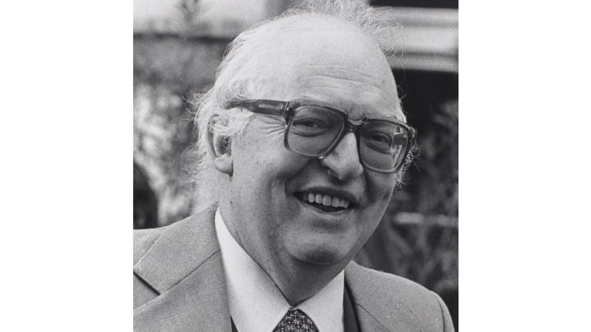Black and white portrait of Friedrich Dürrenmatt. An elderly man smiling in a suit and tie, with white hair and thick glasses, close-up in black and white.