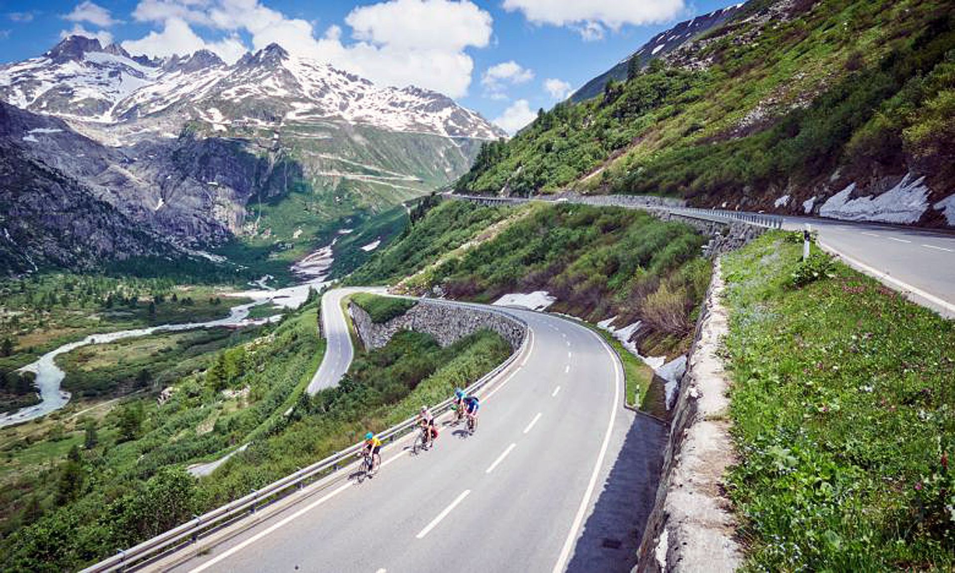 The image displays a serpentine road in a mountainous region with cyclists on it.