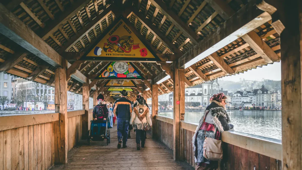 People walking across a covered wooden bridge decorated with colourful panels, some in costume, with water and buildings in the background.