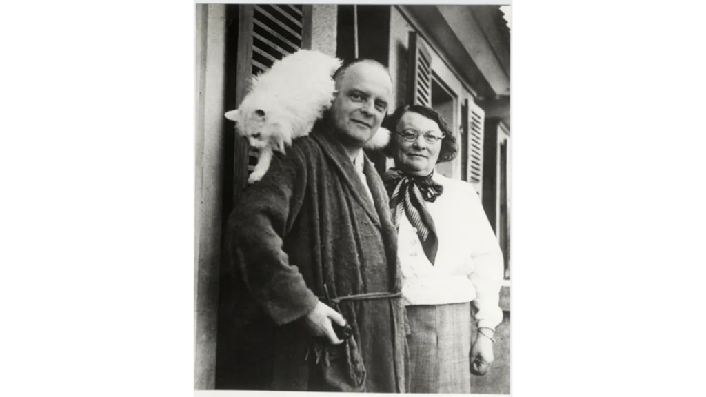 Black and white photograph of a man in a robe with a white cat on his shoulder, standing next to a woman wearing glasses and a scarf.