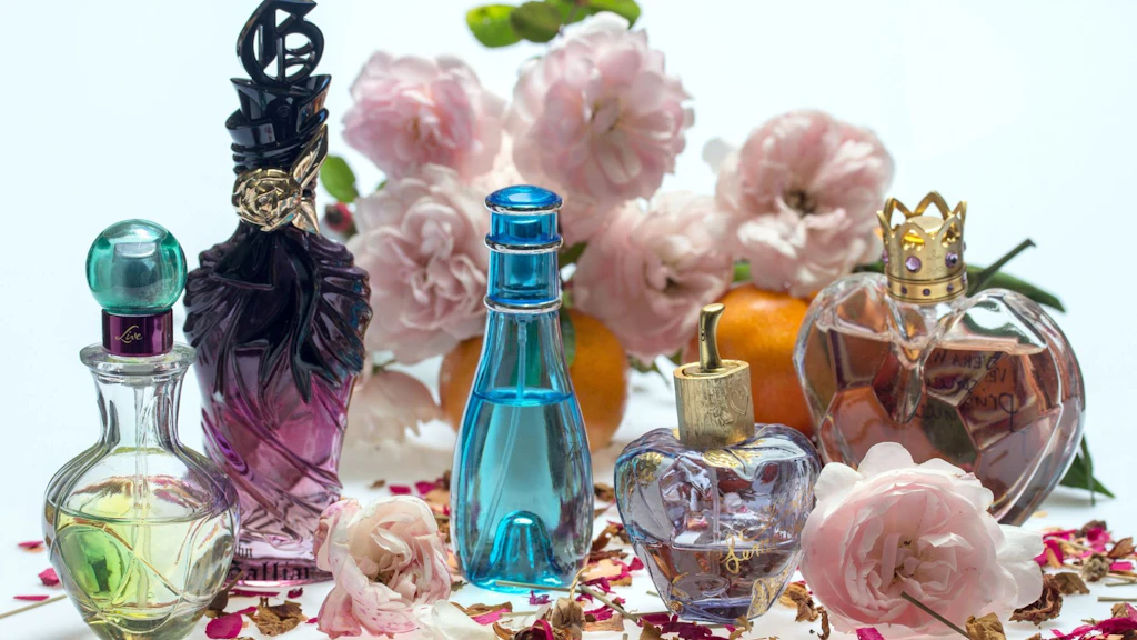 Five perfume bottles of varying shapes and colours arranged on a light surface in front of pale roses, dried petals and a few mandarin oranges.
