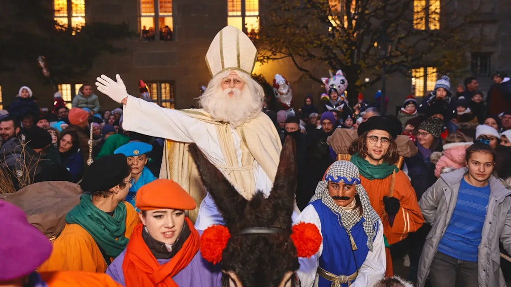 Person dressed as Saint Nicholas greeting the crowd, surrounded by costumed participants during an evening festive parade. 