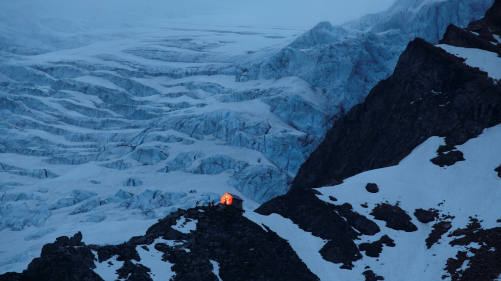 Small illuminated mountain hut on a snowy rocky ridge, with a large blue glacier in the background.