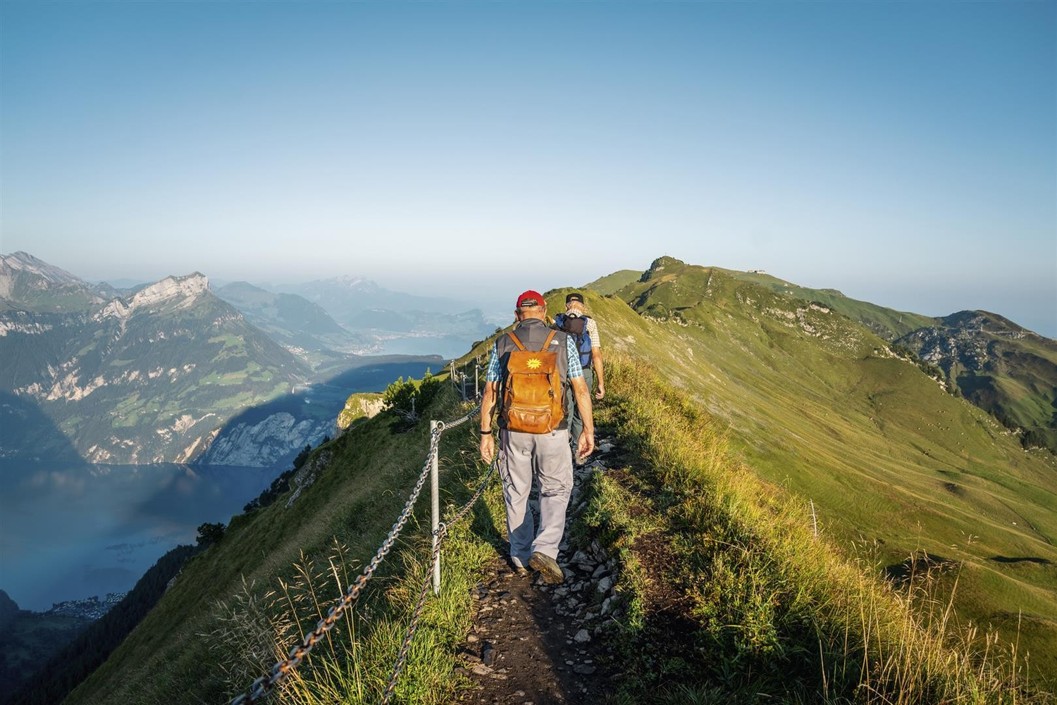 Two hikers walk along a narrow, grassy mountain ridge with a clear blue sky and distant mountains in the background.