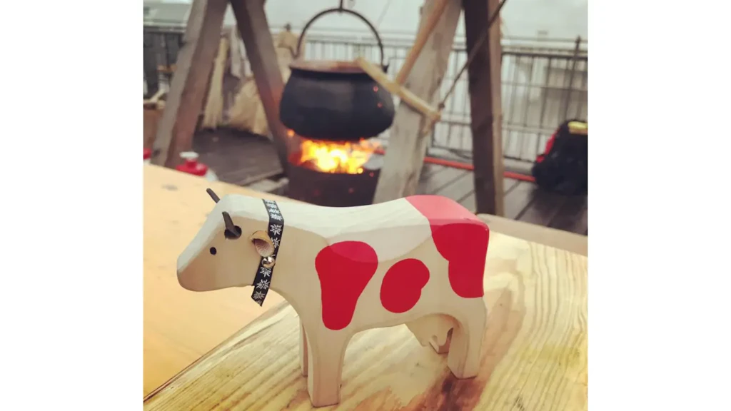 Wooden cow figurine with painted red patches, placed on a wooden surface, wearing a black decorative collar with a small bell.