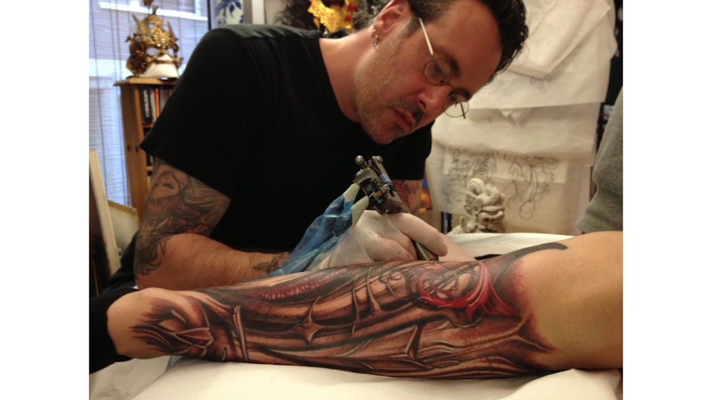 Photograph of a tattoo artist focused on creating a colourful tattoo on a person’s forearm, inside a decorated studio.