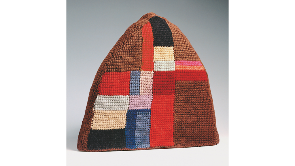 Brown knitted triangular tea cosy decorated with coloured geometric panels (red, blue, cream, pink, black) arranged to form an asymmetrical cross. 