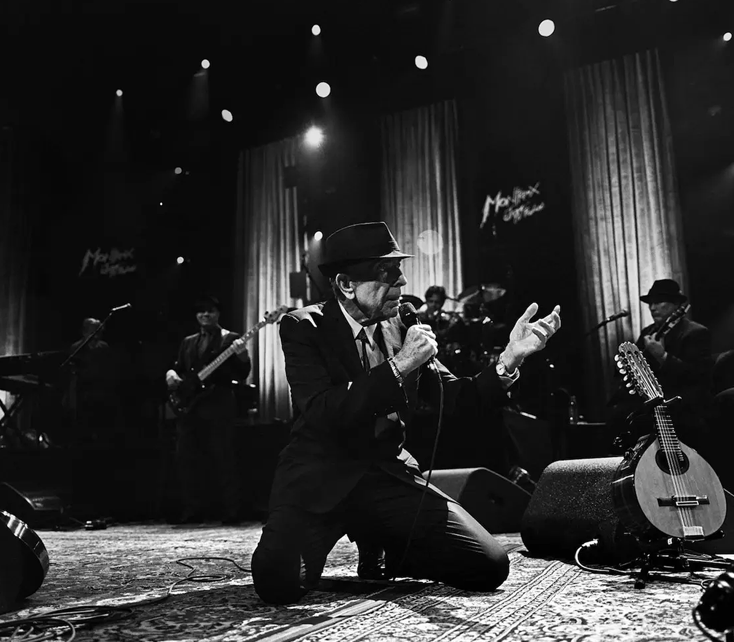 A singer is kneeling on stage, holding a microphone, with musicians playing their instruments in the background.