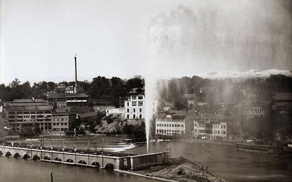 Vintage black-and-white photo of the Jet d’Eau rising from a body of water, with a bridge and buildings on the far shore.