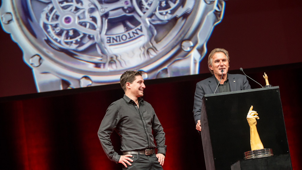Two men stand on stage behind a lectern, a large close-up of a watch mechanism projected behind them and a trophy placed on the lectern.