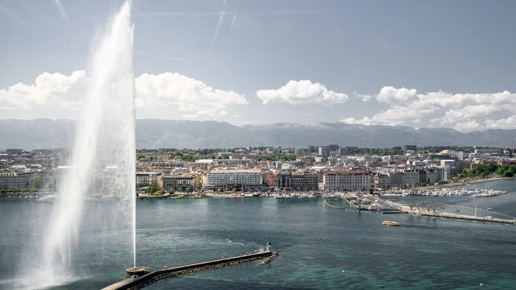 View of Geneva with the Jet d'Eau fountain, Lake Geneva, and the city waterfront under a clear sky.