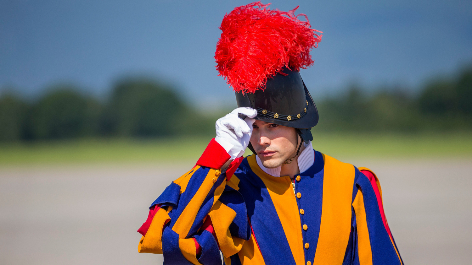 A Swiss Guard in a blue, yellow, and red uniform salutes by touching his black helmet adorned with a red plume, wearing a white glove, outdoors in clear weather.
