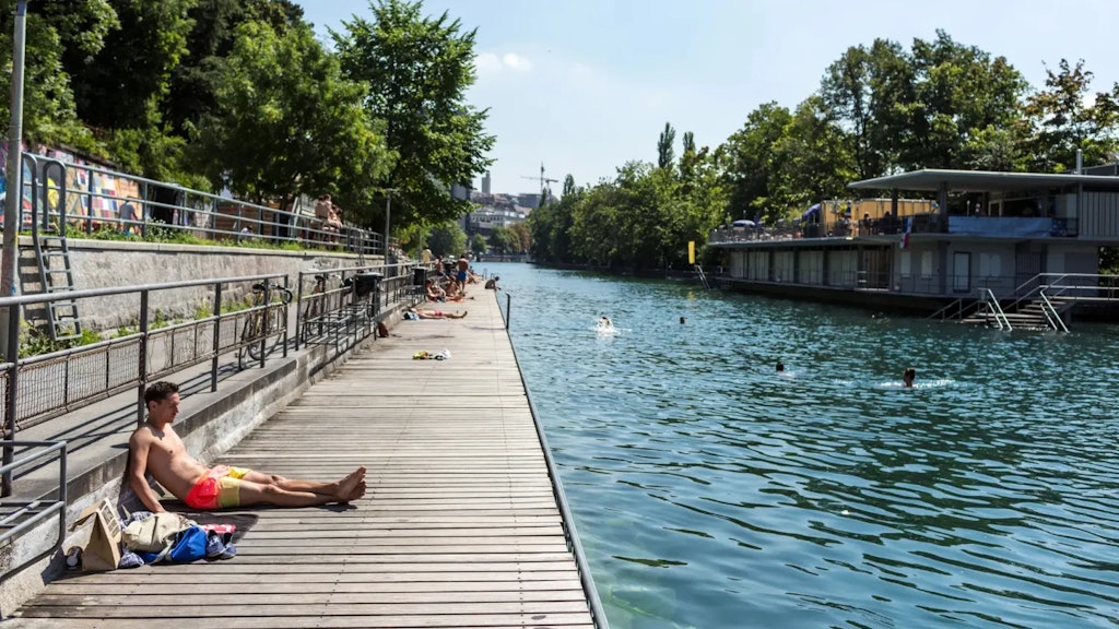 People relax on a wooden deck by a river where others swim, surrounded by trees and buildings under a clear sky.