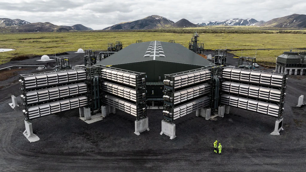 A large industrial plant in Iceland with container-like modules, pipes and fans in a volcanic landscape under a cloudy sky.