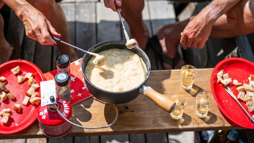 People sharing a cheese fondue outdoors, with pieces of bread and glasses placed on a wooden table.