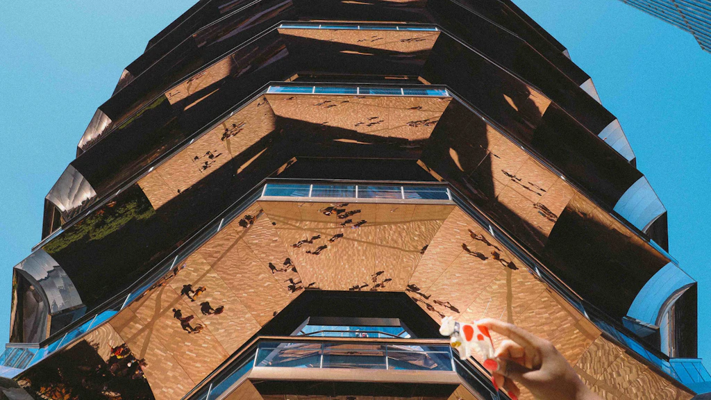 Multi-level architectural structure with reflective surfaces seen from below, with a hand holding a small wooden cow figurine in the foreground.