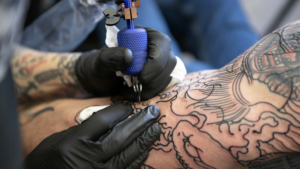 Close-up of a tattoo artist’s gloved hands holding a blue tattoo machine, outlining a black linear pattern on a person’s forearm adorned with other tattoos.