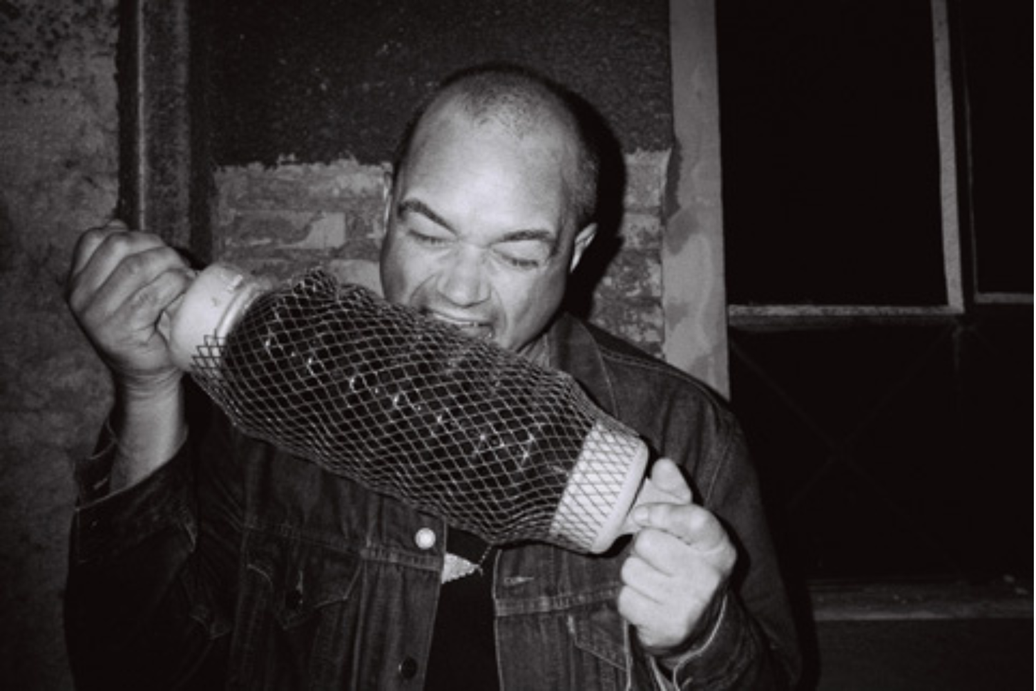 Close-up of a man holding a long mesh-covered cylindrical object, appearing to bite it, in a dimly lit room with rough walls and a partially visible window.