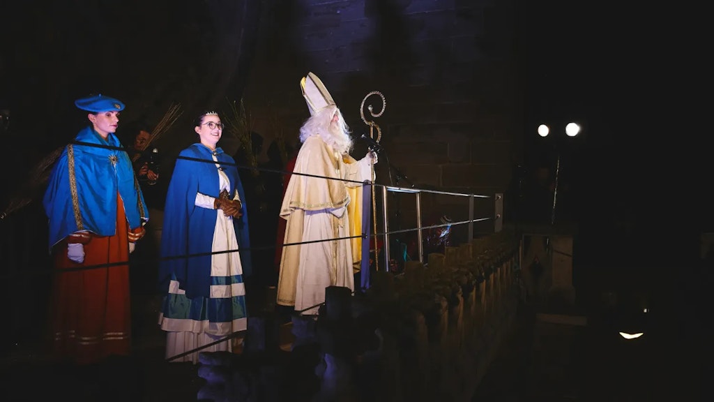 Person dressed as Saint Nicholas holding a bishop’s staff stands on a lit stage at night with two companions in blue costumes.