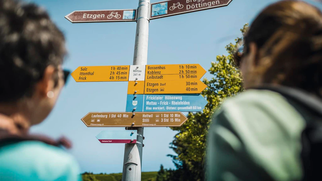 Hiking signpost with coloured directional arrows showing destinations and times on a metal pole, two blurred hikers visible in the foreground.