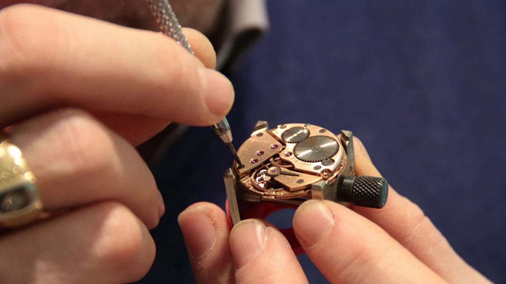 Close-up of metal gears and golden cogwheels from a watch mechanism lying on a dark surface, with a watchmaker’s tool visible.