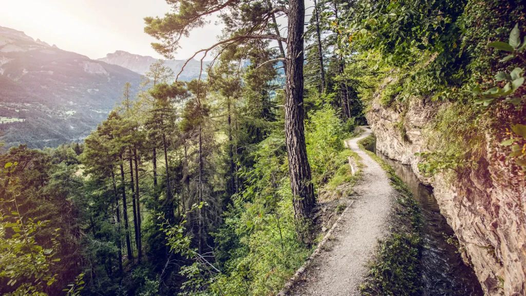 Wooden walkway running beside an irrigation channel on a rocky slope, overlooking a green valley with terraced vineyards under a cloudy sky.