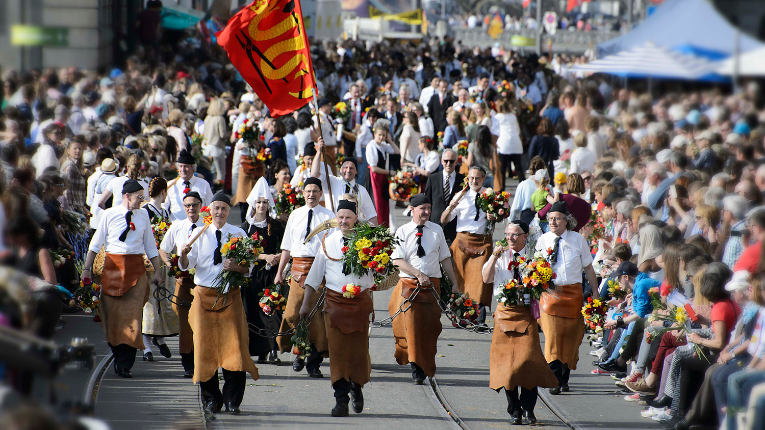 A procession of men in leather aprons and white shirts, carrying flowers and a red flag, marches through a crowded street during an event.
