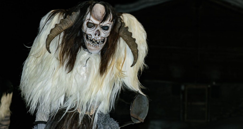 A frightening figure wearing a skull mask, long horns, and a shaggy fur costume stands in a snowy night in front of a dark wooden house.