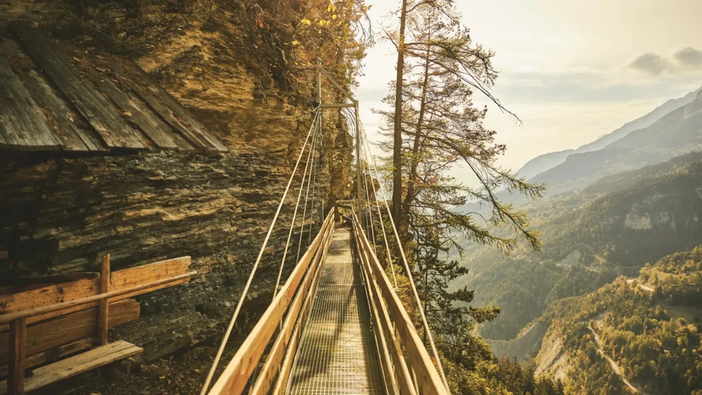 Narrow wooden and metal walkway along a rocky cliff above a mountain landscape, with trees and a downward view over the valley.