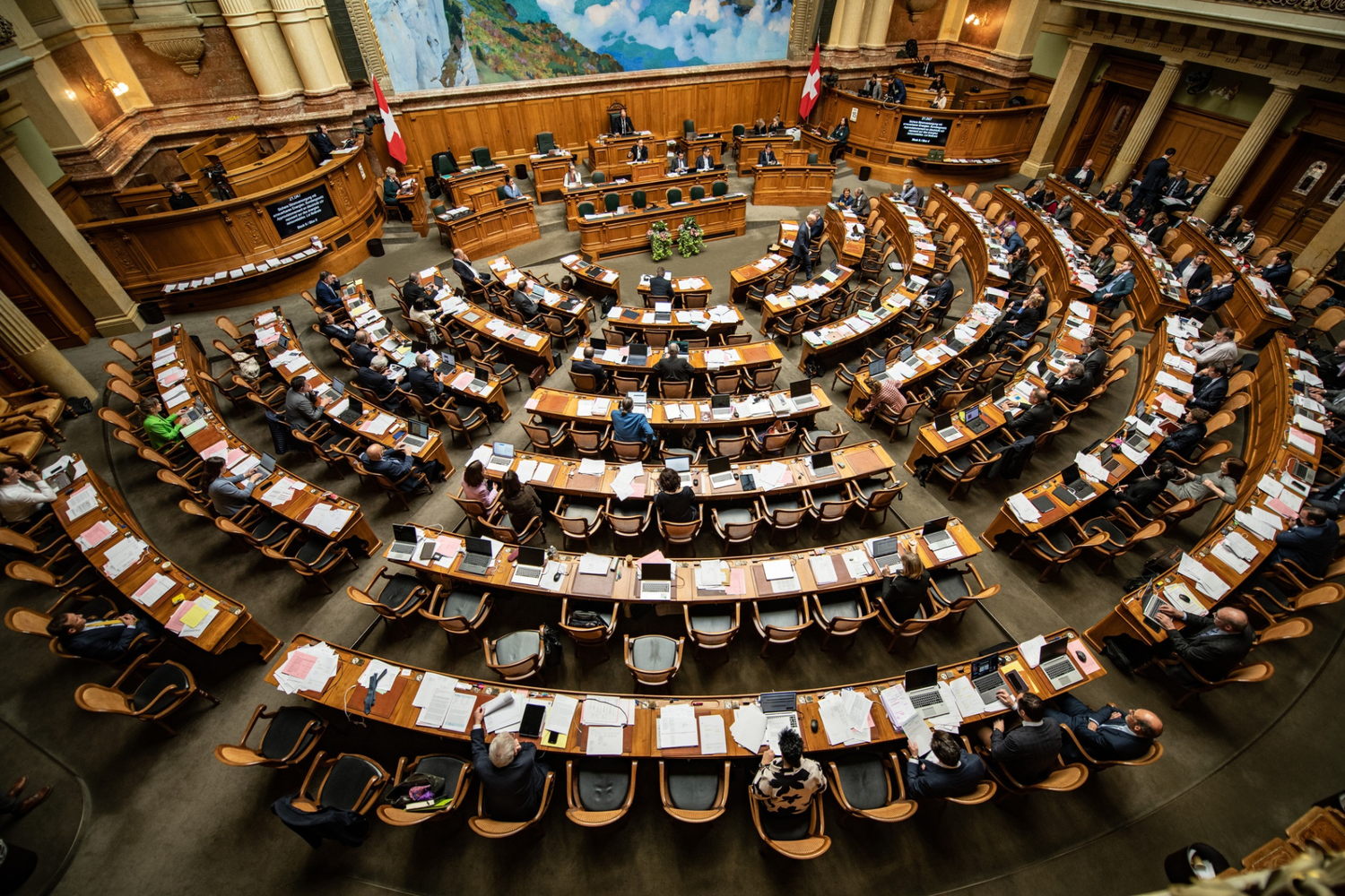 Overhead view of the Swiss National Council: semicircular wooden seating, desks with documents and laptops, Swiss flags visible at the sides.