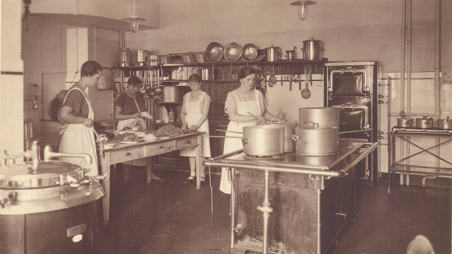 Black and white photo of a 1920s kitchen with four women in aprons preparing food and cooking on a large stove, surrounded by utensils and pots.