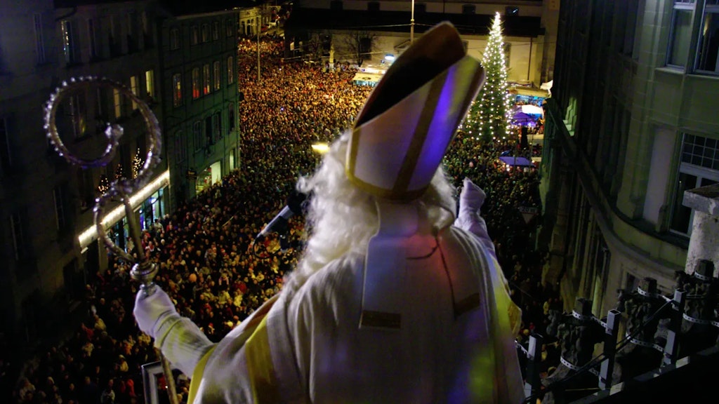 Person dressed as Saint Nicholas holding a bishop’s staff, seen from behind, addressing a large crowd at night before an illuminated Christmas tree. 