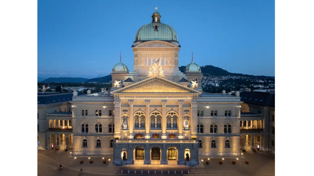 Exterior view of the Federal Palace in Bern at dusk, illuminated neoclassical building with central dome and side wings, hills in the background.