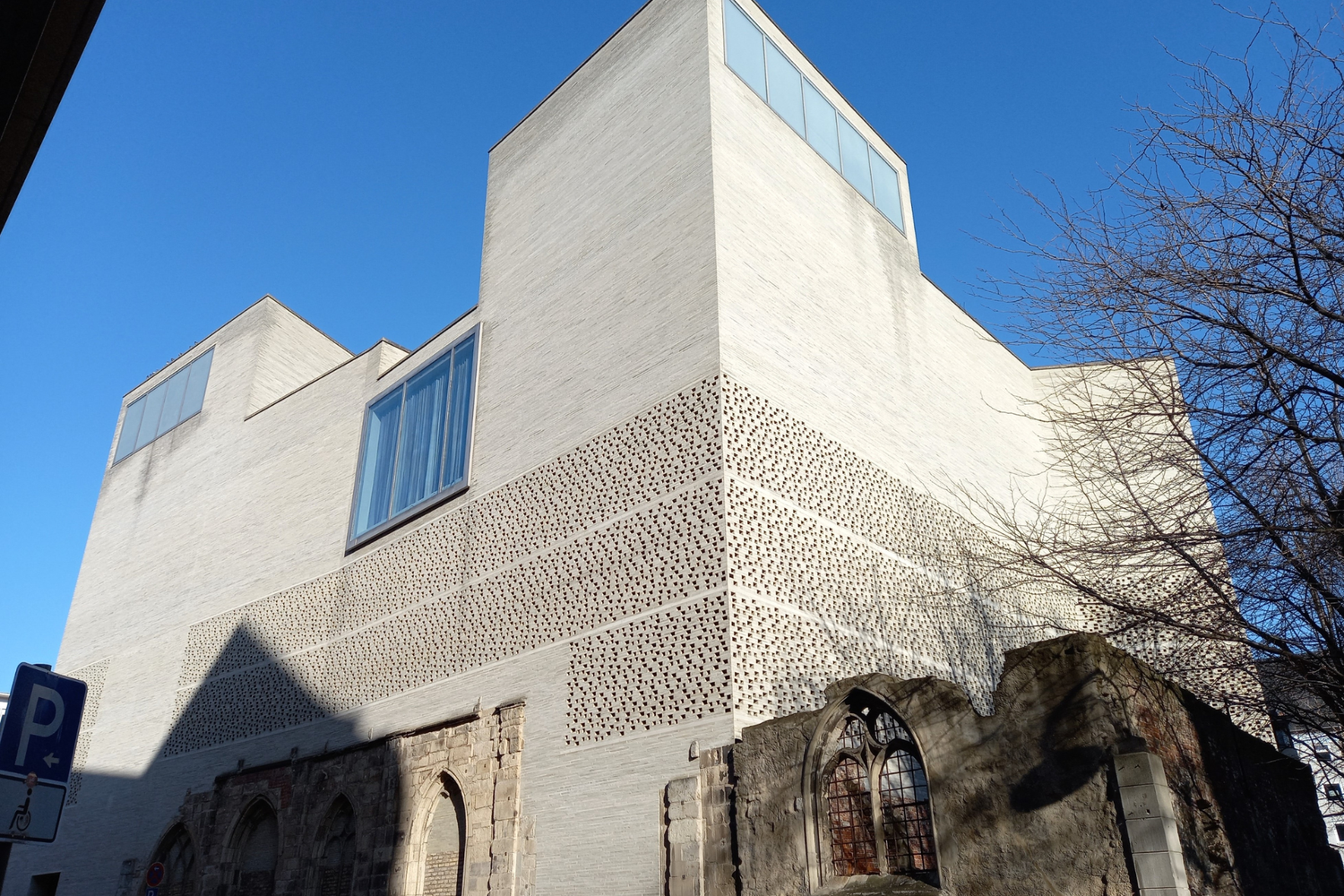 Exterior view of St. Kolumba Church in Cologne, combining Gothic ruins and modern light brick architecture under a clear blue sky.