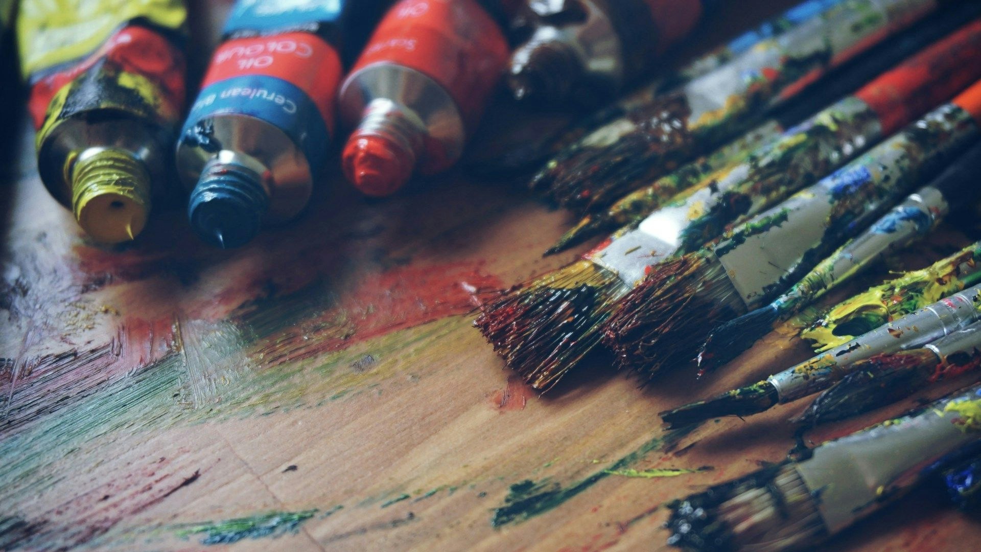 Several open oil paint tubes and paint-splattered brushes resting on a wooden surface covered in colourful paint marks.