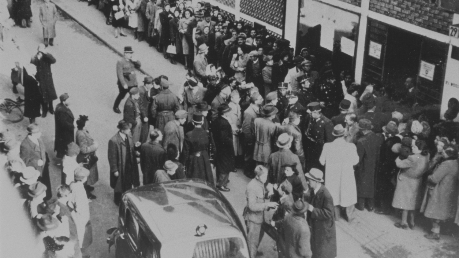 Black and white photo showing a dense crowd of people, some standing in line in front of a building, surrounded by police officers and uniformed men.