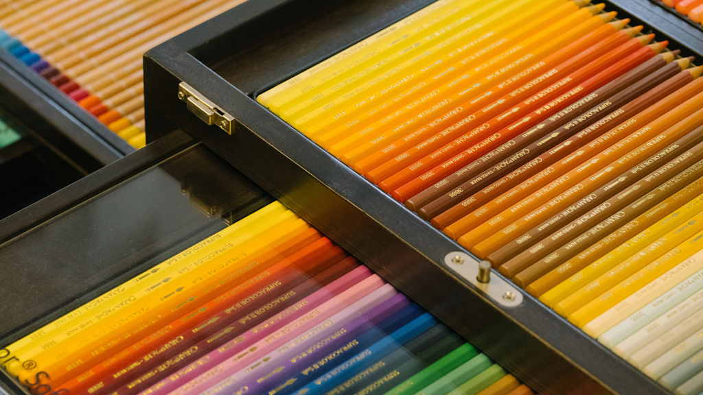 Open wooden box with drawers filled with coloured pencils organised by shade from yellow-orange to violet, viewed from above. 