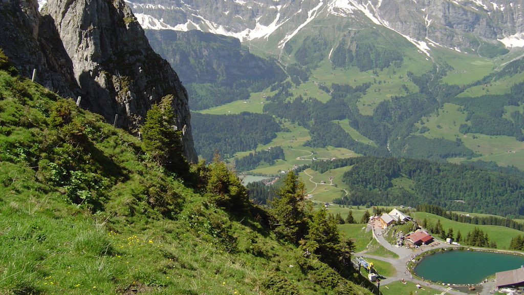Aerial view of an alpine landscape with a green slope, a small lake, and mountain buildings at the base of a rocky cliff.