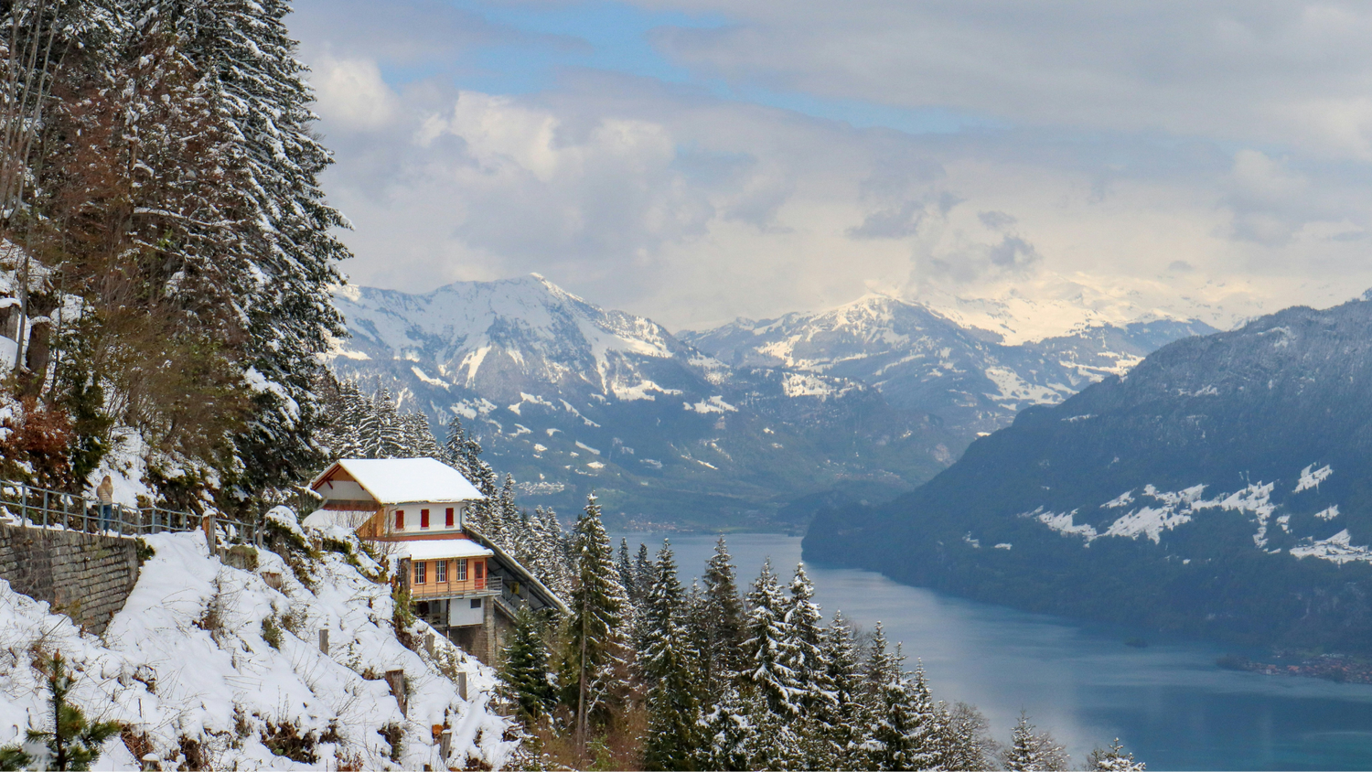 Winter landscape in Interlaken, Switzerland, featuring a mountain house, snow-covered fir trees, a lake, and Alpine peaks under a partly cloudy sky.