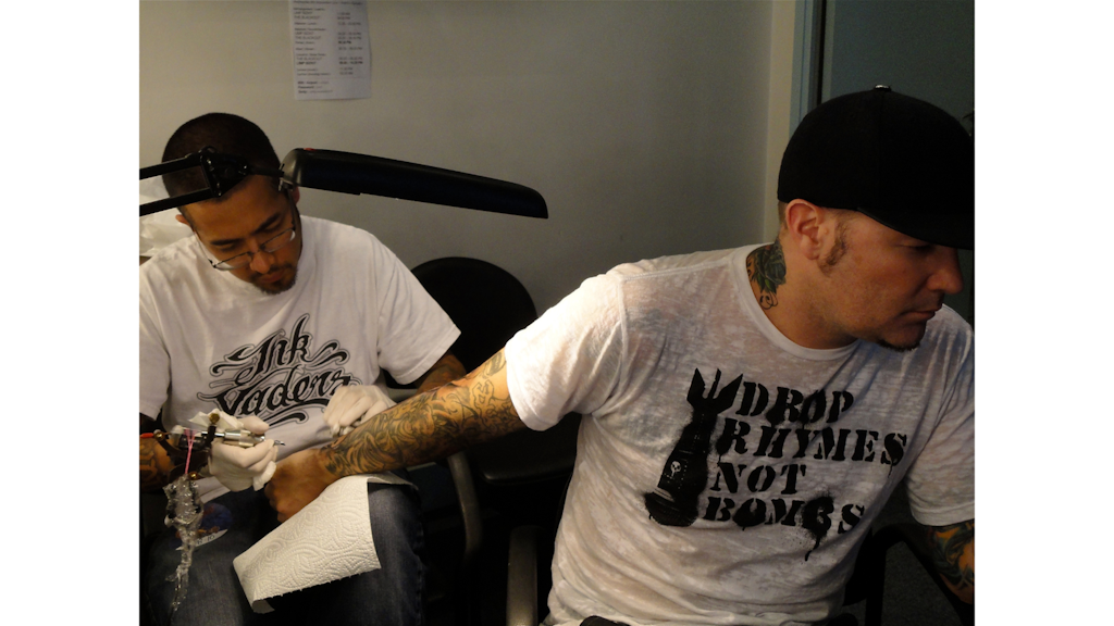 Photograph of a tattoo artist working on the arm of a seated man, both wearing light-coloured T-shirts, inside a tattoo studio.