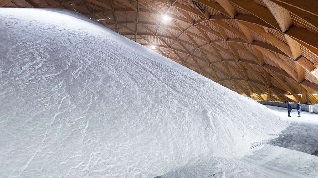Large mound of white salt stored in a spacious covered hall with a wooden roof structure, concrete floor and two people in the distance.