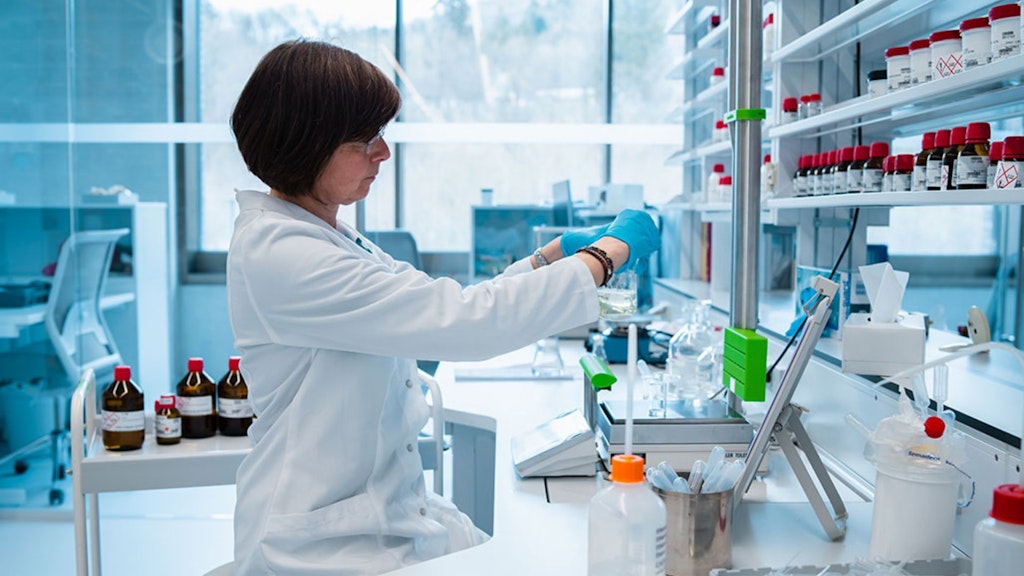 Woman in lab coat and blue gloves handling a beaker on a workbench surrounded by bottles and shelves of chemicals.