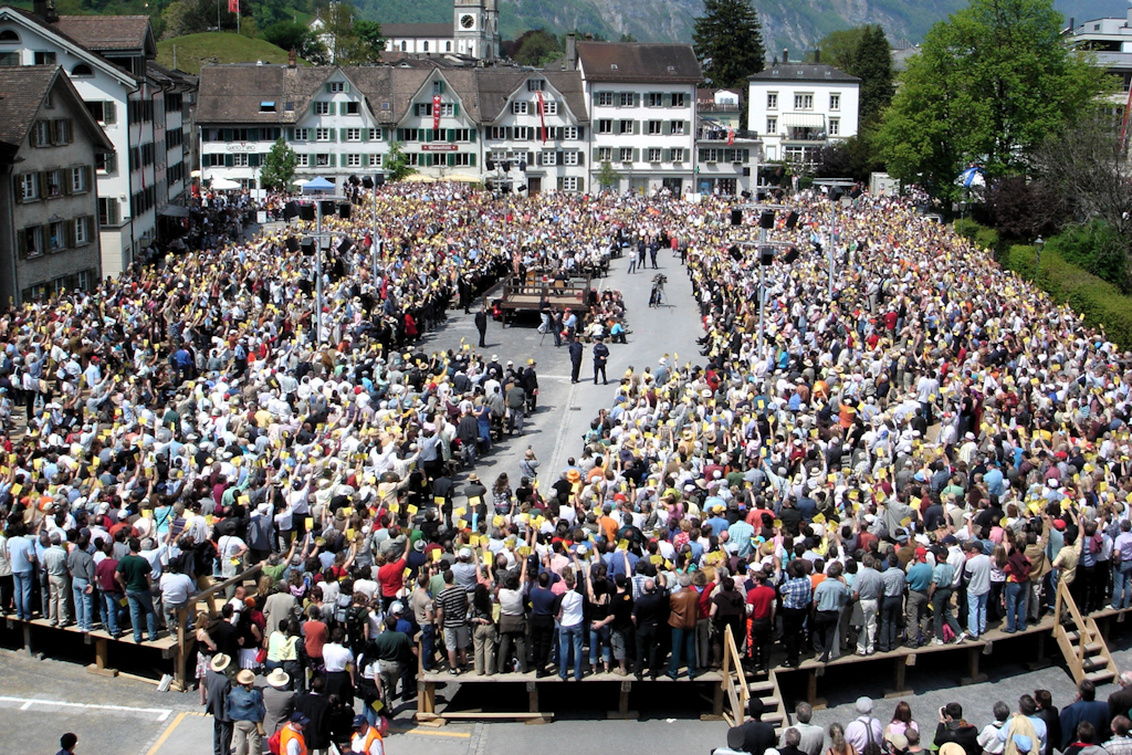 Aerial view of a village square where thousands of people stand in a large circle holding yellow voting cards around a central podium. 